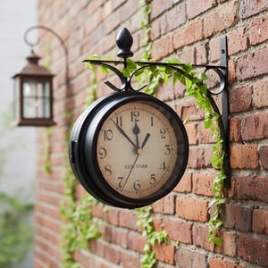 Könnte beinhalten: Eine schwarze, doppelseitige Outdoor-Uhr mit der Aufschrift "NEW YORK" auf dem Zifferblatt, montiert an einer Backsteinmauer. Die Uhr ist von einer dekorativen Metallhalterung und Kletterpflanzen umgeben. Im Hintergrund hängt eine Laterne.