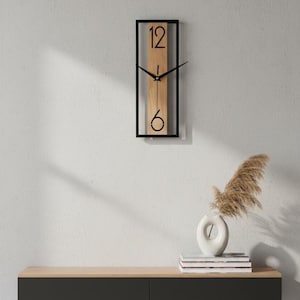 Op de afbeelding: Een rechthoekige wandklok met een zwart frame en een lichtbruin middenstuk. De wijzerplaat heeft zwarte cijfers op de posities 12 en 6, met zwarte wijzers. De klok is aan een witte muur bevestigd. Een witte vaas met gedroogde planten staat op een houten kast.