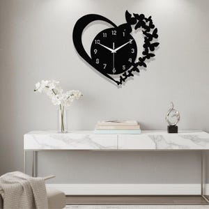 Może przedstawiać: Czarny zegar ścienny w kształcie serca z tarczą i motylkowymi akcentami. Zegar ma białe wskazówki i minimalistyczny design. Zegar jest zamontowany na jasnoszarej ścianie.