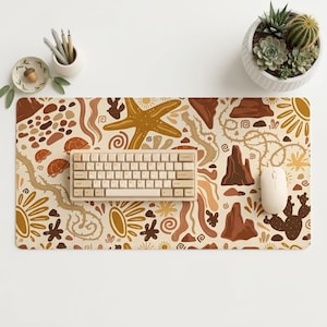 Op de afbeelding: Een rechthoekige bureaumat met een woestijnpatroon in bruin-, beige- en crèmetinten. Het ontwerp bevat illustraties van een zeester, cactussen, schildpadden, zonnen en rotsformaties. Een beige mechanisch toetsenbord en muis liggen op de mat.