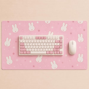May include: A rectangular pink desk mat featuring a repeating pattern of white rabbit heads and small stars. Centered on the mat is a mechanical keyboard with white and light pink keycaps, accompanied by a white computer mouse on the right side.