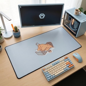 May include: A light blue desk mat with a black border featuring a cartoon cat sleeping on a laptop. A light blue keyboard with orange and beige keys, a light blue mouse, and a computer monitor are also visible.