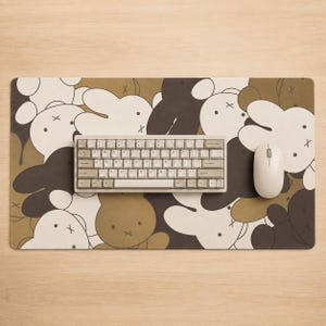 Könnte beinhalten: Eine rechteckige Schreibtischunterlage mit einem sich wiederholenden Muster aus Miffy-Hasenfiguren in Braun-, Creme- und Dunkelgrautönen. Eine kompakte mechanische Tastatur mit hellen Tastenkappen und eine weiße kabellose Computermaus liegen darauf.