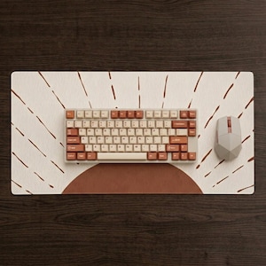 May include: A rectangular desk mat featuring a cream background with a large terracotta semi-circle and radiating brown lines. A mechanical keyboard with cream and terracotta keys sits on the mat, accompanied by a geometric light gray computer mouse.