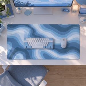 May include: A rectangular desk mat featuring a wavy, abstract pattern in shades of light blue, medium blue, and white. A compact mechanical keyboard with white and light blue keys and a white wireless computer mouse are placed on the mat.