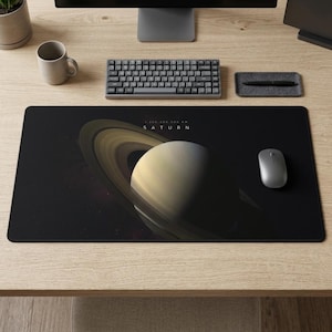 Puede incluir: Una alfombrilla de escritorio rectangular negra con una ilustración de Saturno y sus anillos. La palabra "SATURN" está impresa en blanco. Un ratón inalámbrico, un teclado y un portalápices también están en el escritorio.