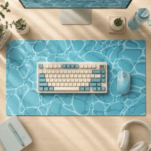 Pode incluir: Um tapete de secretária retangular com um padrão de ondulação de água azul, um teclado mecânico com teclas creme e azuis e um rato de computador azul a condizer. O teclado tem um esquema QWERTY padrão com teclas de cor clara.