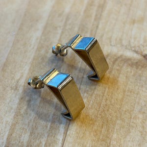 May include: A pair of gold-toned geometric earrings with a simple, modern design. The earrings are made of a thin metal and have a slightly curved shape.