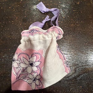 May include: A small white fabric bag with a pink floral print and purple ribbon ties. The bag is made from a handkerchief.