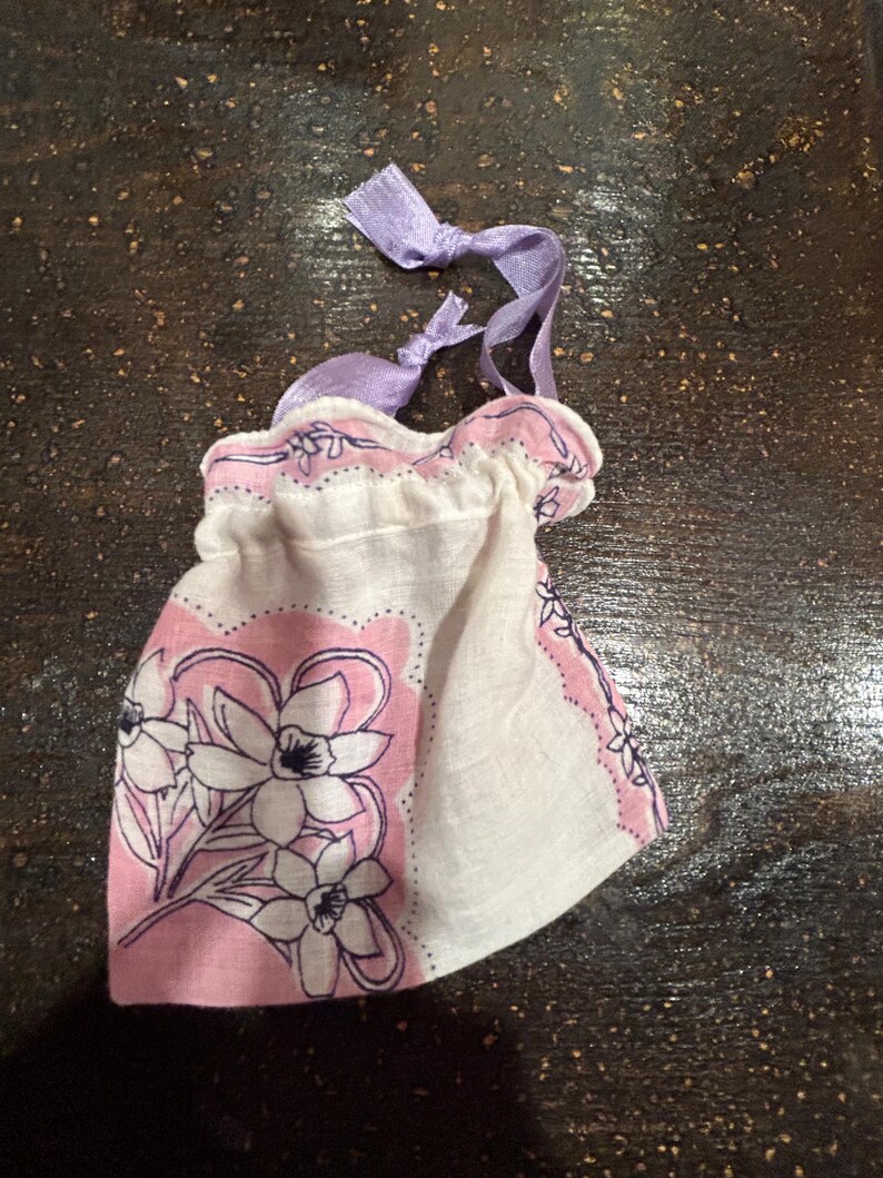May include: A small white fabric bag with a pink floral print and purple ribbon ties. The bag is made from a handkerchief.