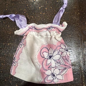 May include: A small white fabric bag with a pink and white floral print and purple ribbon ties. The bag is made from a handkerchief.