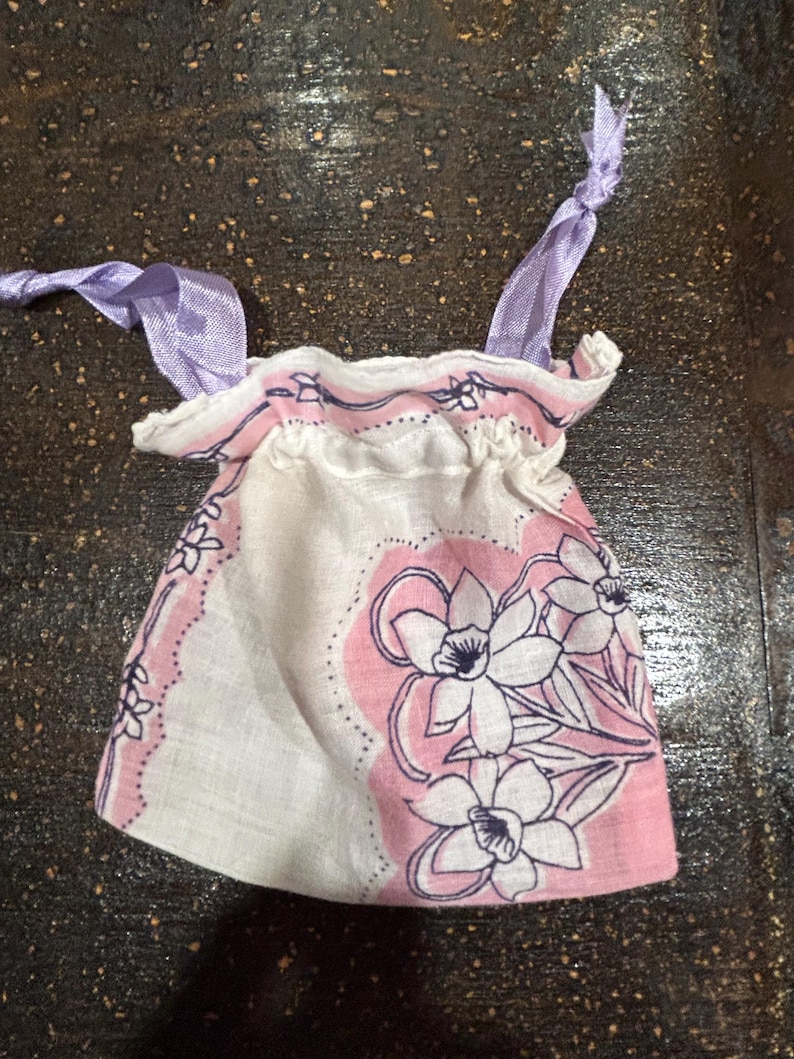 May include: A small white fabric bag with a pink and white floral print and purple ribbon ties. The bag is made from a handkerchief.