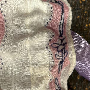 May include: A white fabric with a pink and blue floral pattern. The fabric has a ruffled edge and a purple ribbon attached to it.