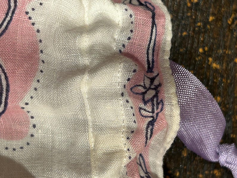 May include: A white fabric with a pink and blue floral pattern. The fabric has a ruffled edge and a purple ribbon attached to it.