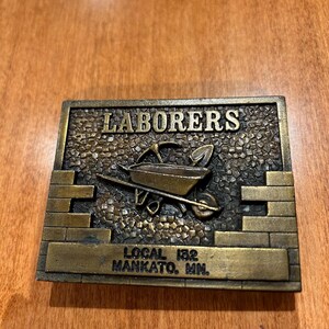 May include: A gold-toned metal belt buckle with a raised design of a brick wall and a wheelbarrow. The text "LABORERS LOCAL 152 MANKATO, MN." is engraved on the buckle.