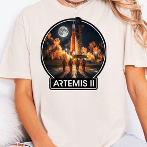 May include: Cream-colored t-shirt featuring a circular graphic of a rocket launch with a full moon in the background. The graphic includes the text "ARTEMIS II" in a white rectangle at the bottom. Four figures in orange suits are walking towards the rocket.