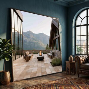 Op de afbeelding: Een grote rechthoekige vloerspiegel met een dun donker frame die een buitenterras weerspiegelt. De reflectie toont een stenen terras met een vuurplaats, buitenmeubilair en uitzicht op bergen onder een heldere lucht in een blauwe kamer.