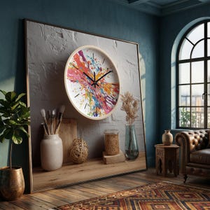May include: A round wall clock with a light wood frame and a colorful abstract splatter design. The clock is mounted on a textured gray wall. The clock hands are black.