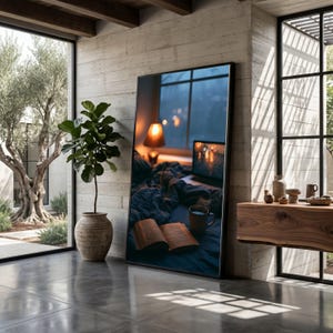 May include: A large rectangular floor mirror with a black frame reflecting a cozy bedroom scene featuring an open book, a dark mug, a laptop, and a glowing lamp on a bed, positioned next to a potted fiddle leaf fig plant in a textured ceramic pot.