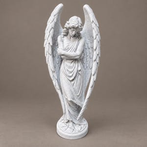 May include: A light gray stone-textured angel statue featuring large, detailed wings extending upward. The figure has long hair, a draped robe, and arms crossed over the chest, standing on a circular base with a weathered, speckled finish.