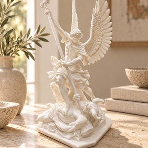 May include: A white sculpture depicting an angel with large feathered wings standing over a horned figure. The angel holds a long staff topped with a cross, wearing draped clothing. The figure is positioned on a flat, angular base.