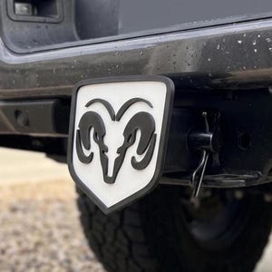 May include: A black and white Dodge Ram logo hitch cover. The hitch cover features a white background with a black Ram logo and a black border. The hitch cover is attached to a black vehicle.