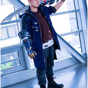 Peut inclure: Une personne portant un costume de cosplay inspiré du jeu vidéo Devil May Cry. La tenue comprend un long manteau bleu foncé, une chemise marron et un jean foncé. Le costume comprend également un bras mécanique et une grande épée.