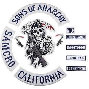 Op de afbeelding: Een verzameling wit en marineblauw geborduurde patches. De centrale patch toont een zeisdrager met een schedelgezicht die een geweer en een zeis vasthoudt. De patches bevatten de woorden "SONS OF ANARCHY", "SAMCRO", "CALIFORNIA", "MC", "MEN OF MAYHEM", "REDWOOD", "ORIGINAL" en "PRESIDENT".