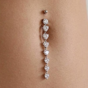 Può includere: Un piercing all'ombelico argentato con un design pendente di pietre preziose chiare a forma di cuore. Il gioiello è mostrato su uno sfondo colorato, con un top bianco visibile nella parte superiore dell'immagine.
