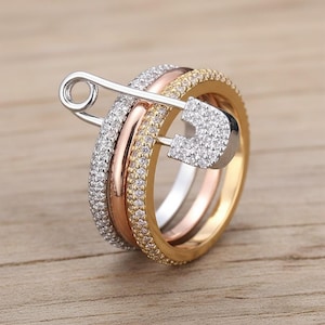 Könnte beinhalten: Ein Set aus drei Ringen in Silber-, Roségold- und Goldtönen, verbunden mit einer silbernen Sicherheitsnadel. Die Ringe sind mit kleinen, funkelnden Steinen besetzt, was ein glamouröses und auffälliges Design ergibt. Die Sicherheitsnadel verleiht einen einzigartigen, modernen Touch.