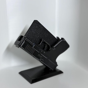 May include: A black 3D-printed handgun display stand. The stand is designed to hold a handgun at an angle, with a textured back and a rectangular base. The handgun is black and appears to be a replica.