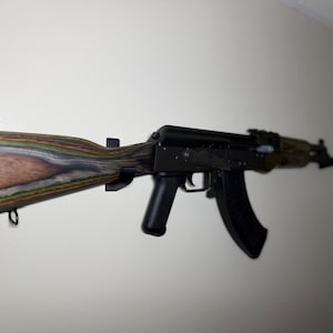 May include: An AK-47 style rifle with a black body and a multi-coloured wooden stock. The stock features shades of brown, green, and grey. The rifle is mounted on a white wall.