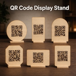 May include: Six light-coloured wooden QR code display stands in various shapes including circular, rectangular, arched, and octagonal. Each stand features a dark brown engraved QR code on the front surface and is supported by a small wooden base.