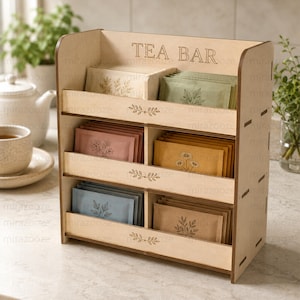 Può includere: Un organizer per tè in legno chiaro a tre livelli e sei scomparti. Il pannello superiore presenta il testo inciso TEA BAR. La parte anteriore di ogni ripiano è decorata con un piccolo motivo a foglie. L'organizer è riempito con varie bustine di tè.