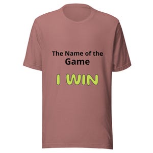 Puede incluir: Una camiseta de color rosa polvoriento de manga corta. El texto "The Name of the Game" está en negro, y "I WIN" en verde brillante con un contorno amarillo. La camiseta está hecha de un material suave.