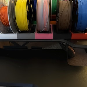 May include: A multi-tiered storage rack holding numerous circular spools of 3D printer filament in various colors including orange, yellow, blue, pink, green, and brown. The spools are organized in individual slots on a black frame structure.