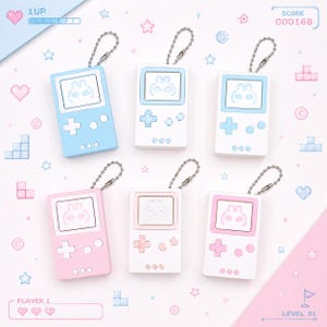 Puede incluir: Seis llaveros de consola de juegos portátil dispuestos en dos filas. Cada uno cuenta con una pantalla con un gráfico de conejo, una cruceta, dos botones circulares y tres puntos pequeños. Los colores incluyen combinaciones de azul claro, blanco y rosa pastel.