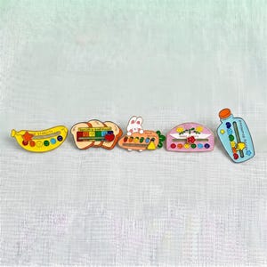 May include: Five colorful enamel pins featuring sliding elements. The pins include a yellow banana with 'and stability', a bread slice with 'banish a bad mood', a rabbit with 'social battery', a pink arch with 'keep happy', and a blue bottle with 'mood download'.