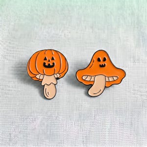 Pode incluir: Dois pins de esmalte com designs de cogumelos laranja. O pin da esquerda é um cogumelo em forma de abóbora com um rosto de jack-o'-lantern. O pin da direita é um cogumelo em forma de fantasma com um rosto simples, ambos com contornos pretos.