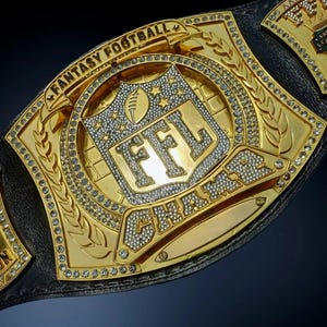 Puede incluir: Un cinturón de campeón de Fantasy Football dorado con las letras "FFL" en el centro. El cinturón está adornado con pedrería y las palabras "Fantasy Football Champion". El cinturón está sobre una correa de cuero negro.