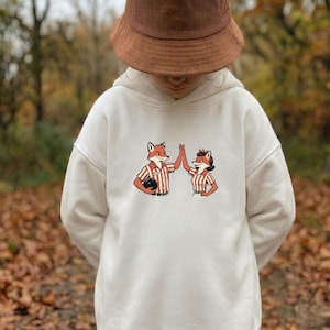 May include: Cream-colored hoodie featuring a cartoon illustration of two foxes giving a high-five. The fox on the left is holding a bowling ball and wearing a red and white striped shirt. The fox on the right is wearing a matching shirt. The person in the image is wearing a brown corduroy bucket hat.