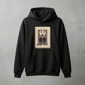 Puede incluir: Sudadera con capucha negra con un gran gráfico de un enchufe eléctrico. El diseño del enchufe presenta un túnel con coches que lo atraviesan. El gráfico es en tonos de marrón, beige y gris.