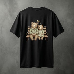 May include: Black t-shirt featuring a whimsical illustration of two teddy bears seated on a park bench. The bears are dressed in vintage-style clothing, with one wearing a green jacket and the other a light green dress. A small bird perches above them.