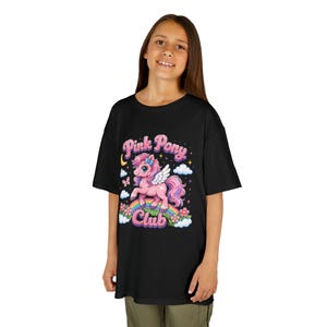 May include: A black t-shirt featuring a graphic print of a pink winged pony standing on a rainbow. The design includes the text Pink Pony Club in a glittery pink font, surrounded by white clouds, small flowers, a yellow crescent moon, and stars.