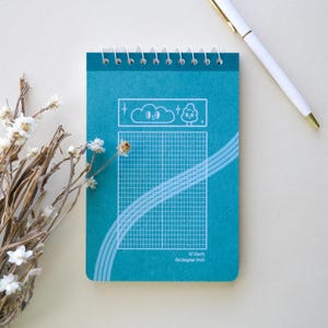 May include: A teal spiral-bound notepad with a grid pattern and cloud and tree illustrations. The notepad has a white spiral binding and a white pen is next to it. The notepad has the text "40 Sheets Rectangular Grids".