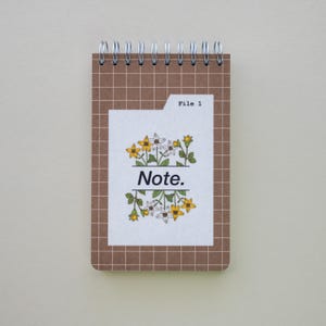 May include: A small spiral-bound notepad with a brown grid-patterned cover. The notepad features a white label with the word "Note." and the text "File 1." above a floral design with yellow and white flowers.