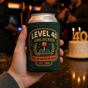 May include: A dark green foam can cooler featuring a pixel art graphic of a joystick and the text LEVEL 40 UNLOCKED, STILL NO INSTRUCTION MANUAL, and EST. 1986. The design includes orange and yellow accents, laurel branches, and a banner.