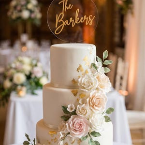 May include: A three-tiered white cake decorated with gold leaf flakes and a cascade of white, cream, and pale pink sugar roses with green leaves. A circular clear acrylic cake topper features the gold script text The Barkers on top.