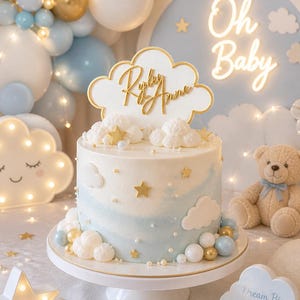 Puede incluir: Una tarta de dos pisos con glaseado azul y blanco, decorada con detalles de nubes y estrellas. Un adorno para tarta con forma de nube y borde dorado que dice "Riley Anne". La tarta está sobre un pedestal blanco, rodeada de globos y un oso de peluche.
