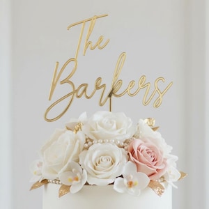 Puede incluir: Una tarta de boda blanca coronada con un adorno dorado que dice "The Barkers" en escritura cursiva. La tarta está decorada con rosas blancas y rosas, perlas y detalles de pan de oro.